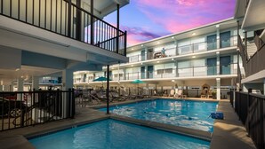 Una piscina con sillas de descanso y sombrillas, rodeada de edificios de varios pisos con balcones.