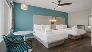 Habitación de hotel con cama, dos mesitas de noche, ventilador de techo, lámpara de pared, ventana con cortinas y una pequeña mesa redonda con una silla.