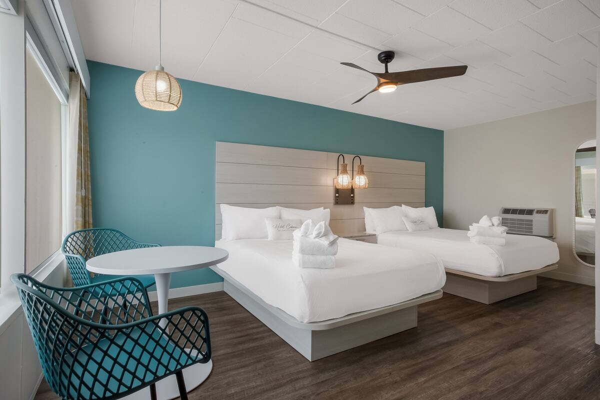 A hotel room with a bed, two bedside tables, a ceiling fan, a wall-mounted light fixture, a window with curtains, and a small round table with a chair.