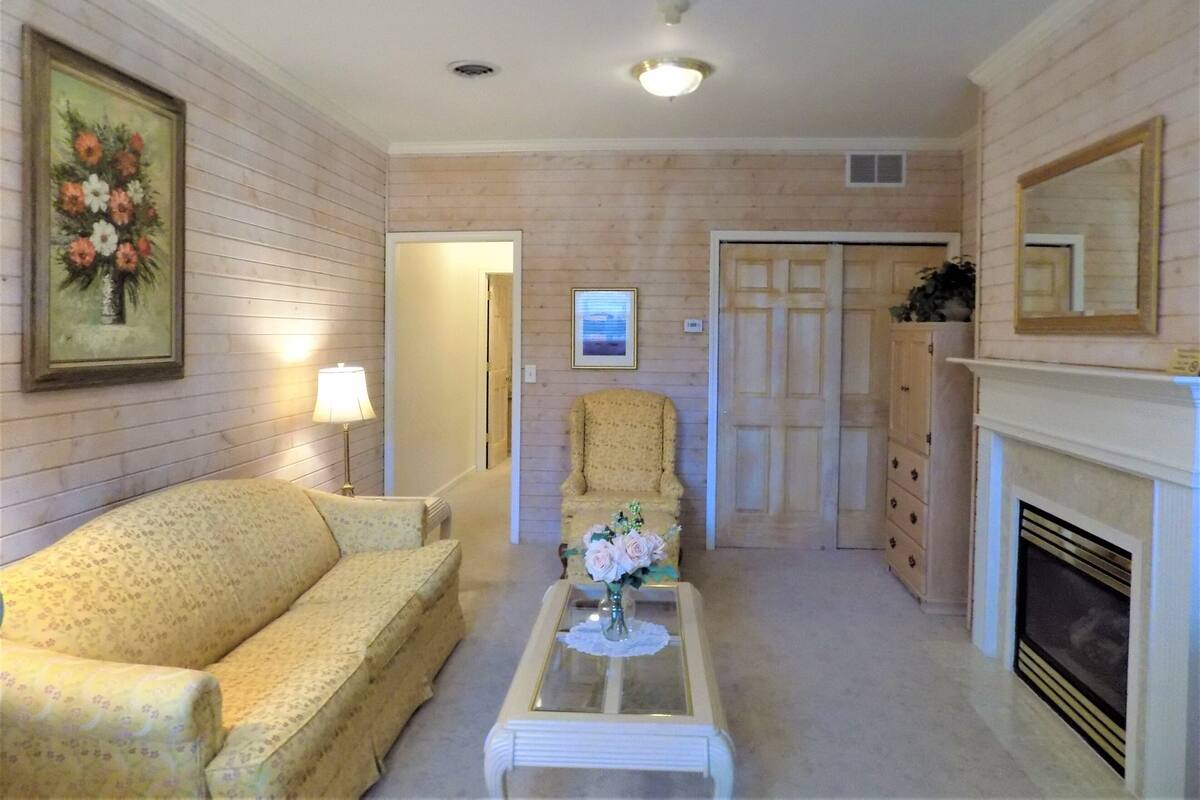 Una sala de estar con sofá, una silla, una mesa de centro con un jarrón, chimenea y una puerta que da a otra habitación.
