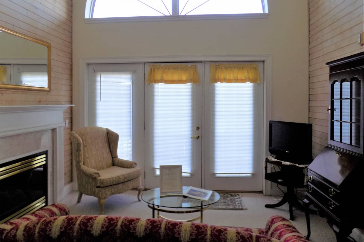 Sala de estar con chimenea, televisor, sofá y sillón. Se observan dos ventanas con cortinas y un tragaluz arriba.