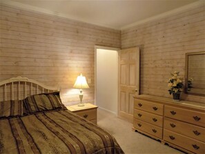 A bedroom with a wooden paneled wall, a bed with a striped bedspread, a bedside lamp, a dresser with drawers, and a mirror on the wall. - The Rukmapura Park Hotel (Fairfield)