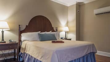 Una cama bien hecha con cabecera de madera, dos mesitas de noche con lámparas, una mesita auxiliar y un aire acondicionado instalado en la pared.