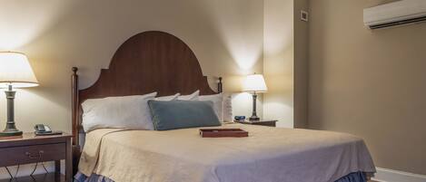 A neatly made bed with a wooden headboard, two bedside tables with lamps, a nightstand, and a wall-mounted air conditioner.