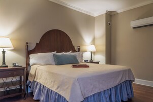 Una cama bien hecha con cabecera de madera, dos mesitas de noche con lámparas, una mesita auxiliar y un aire acondicionado instalado en la pared.