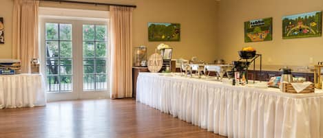 A banquet hall with a long table set for a meal, a buffet area, and a view of the outdoors through large windows.