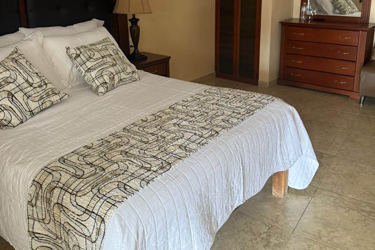 A neatly made bed with a patterned bedspread, a bedside lamp, and a mirror on the wall.