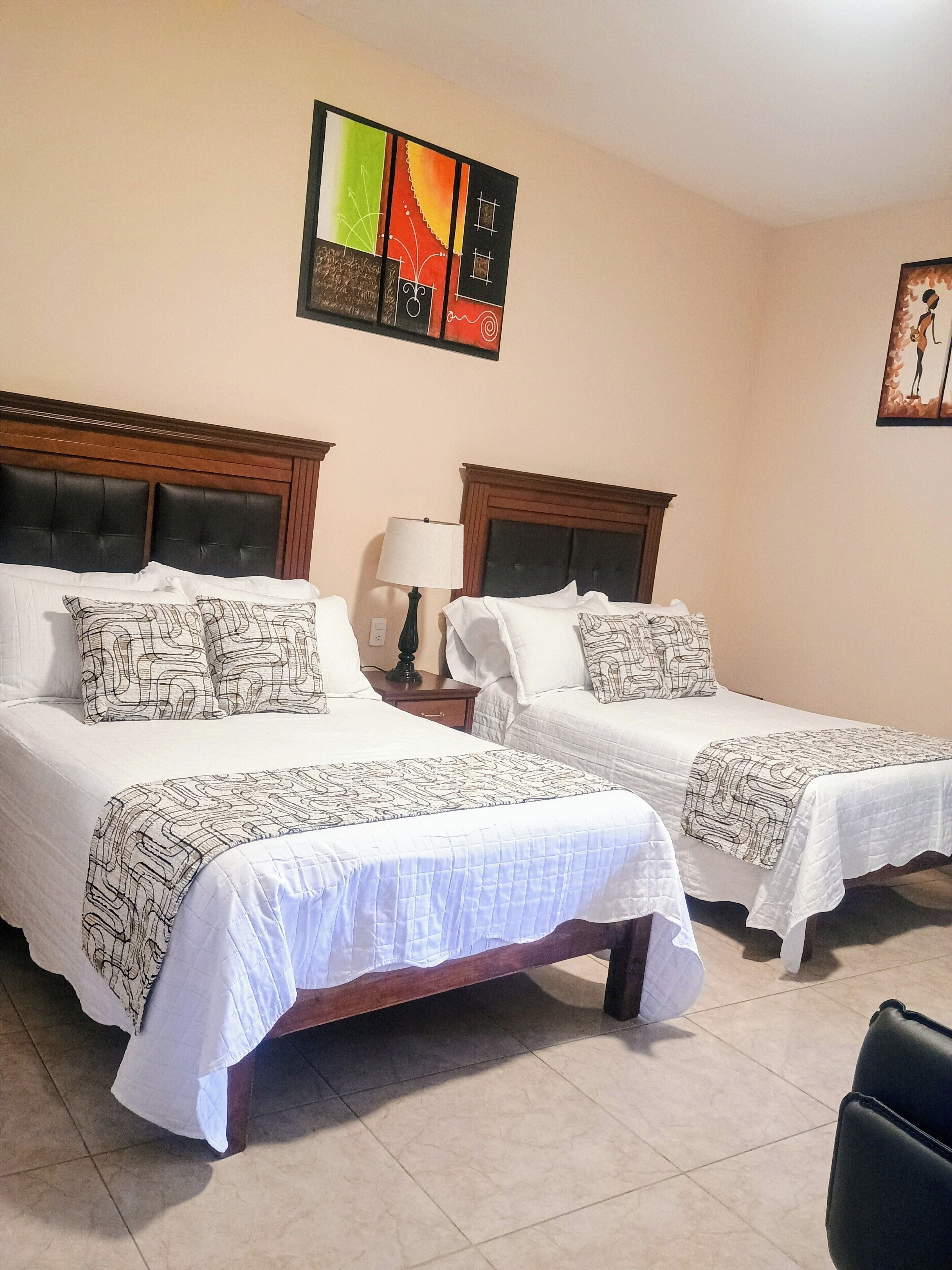 A hotel room with two beds, each with a headboard and a bedside table with a lamp.