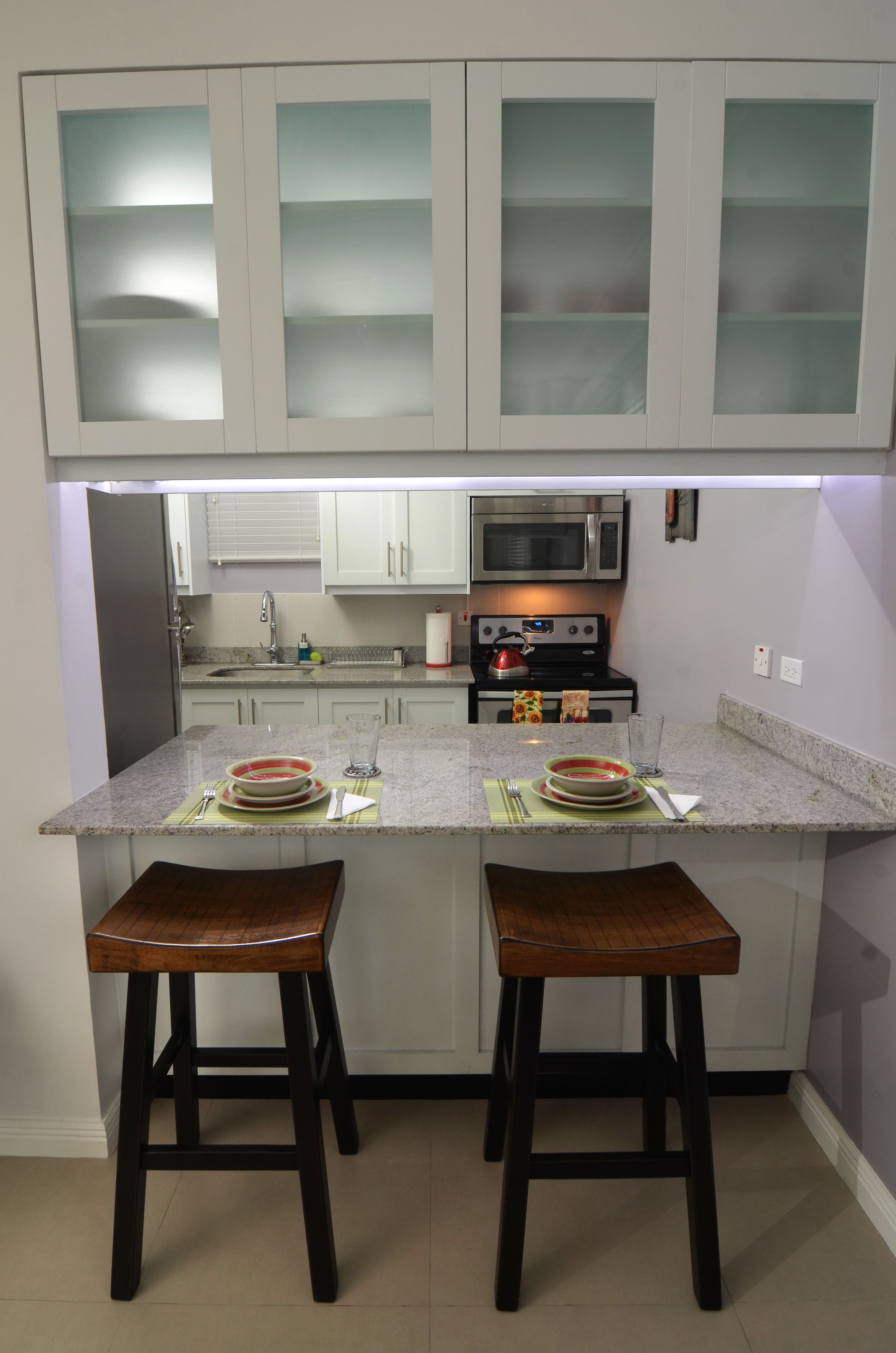 A kitchen with white cabinets, a stainless steel refrigerator, a microwave, and a stove. There are two wooden stools and a glass on the counter.