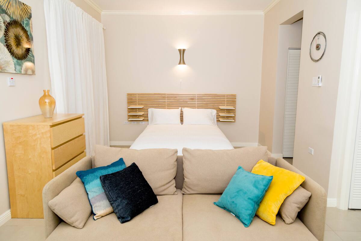 Une chambre d’hôtel avec un lit, une commode en bois, un canapé beige agrémenté de coussins colorés et un luminaire fixé au mur.