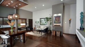 A modern living room with a wooden dining table, dark sofas, and a large window with blinds. - New Kingston Apartment at The Palms (Kingston)