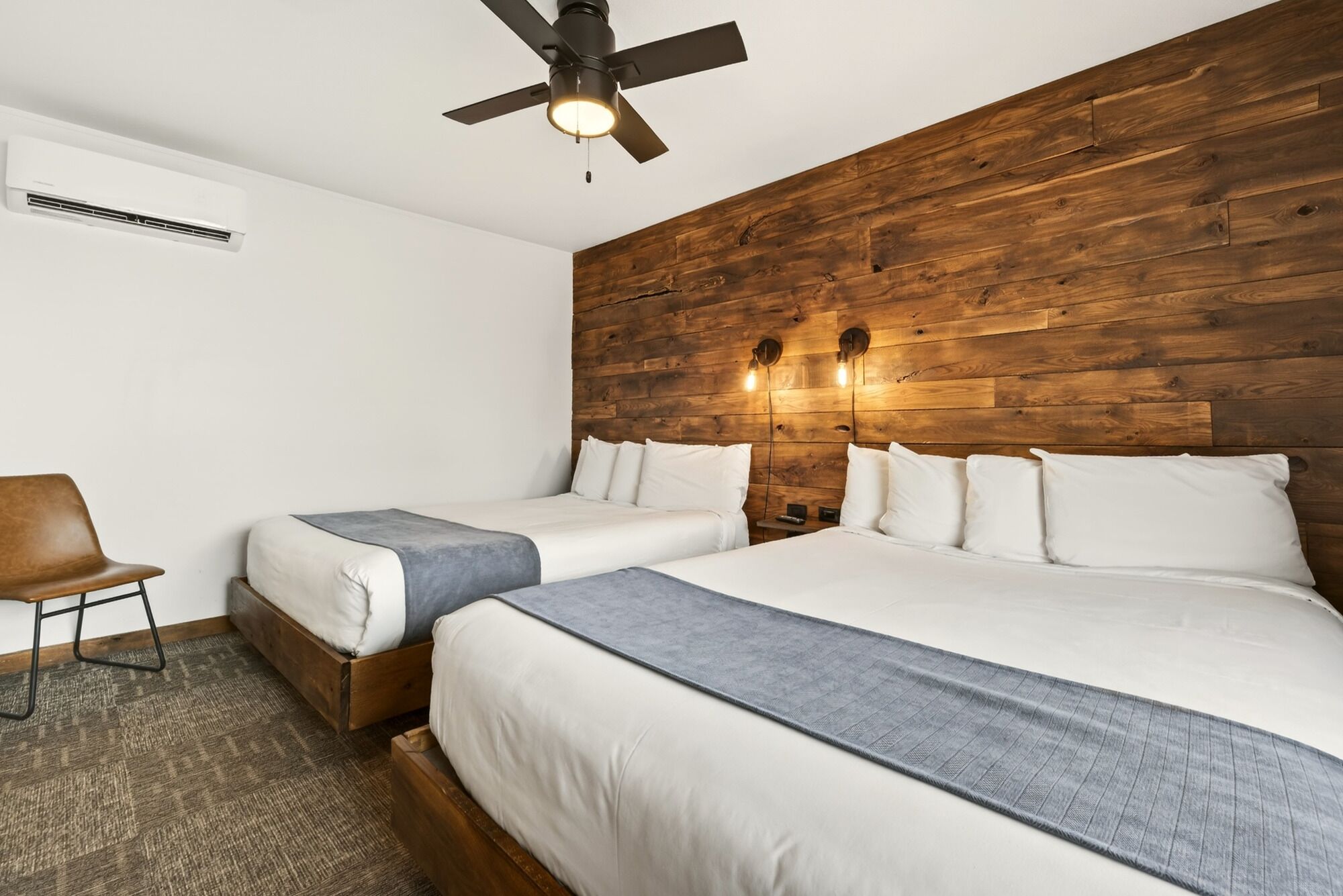 A hotel room with two beds, a ceiling fan, a wooden accent wall, and a chair.