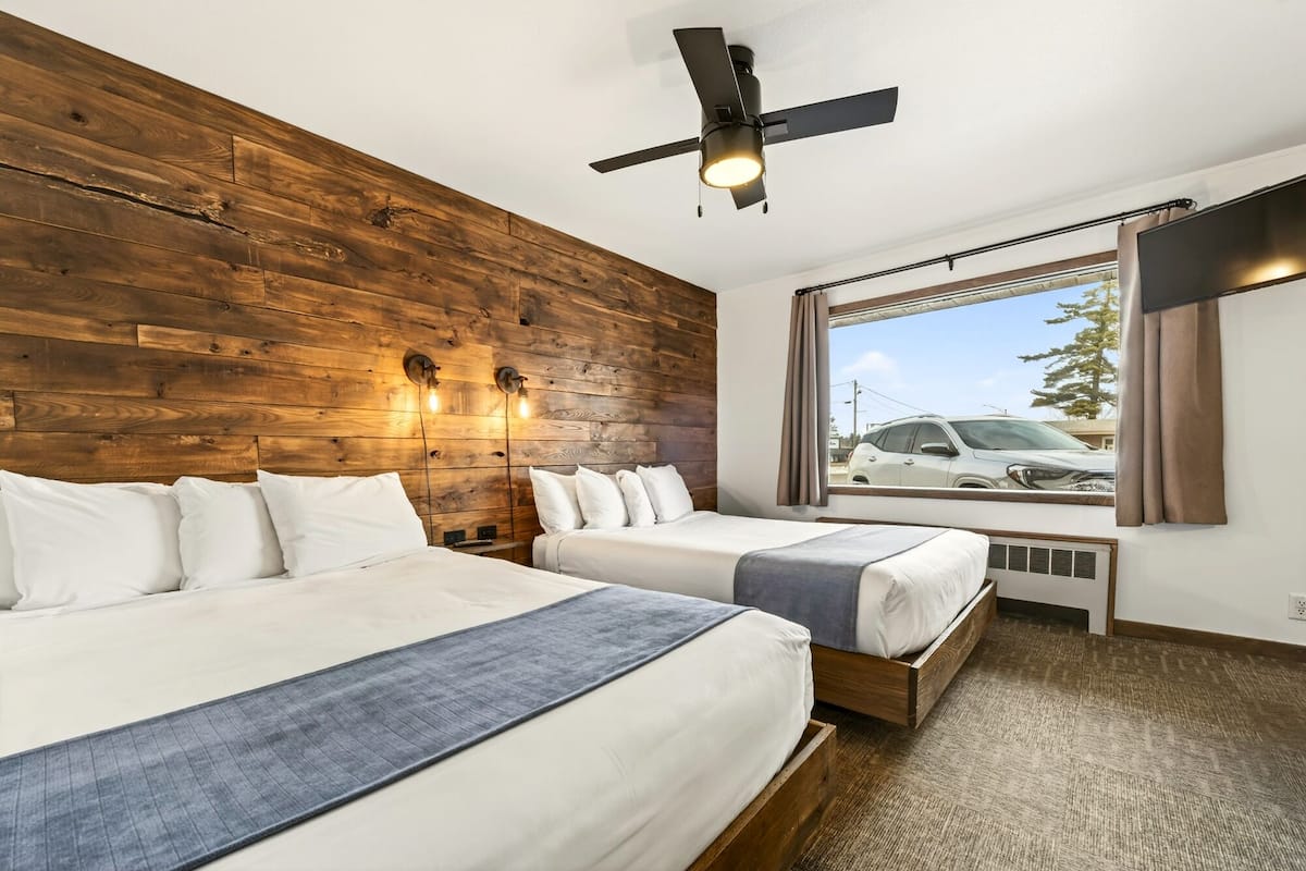 A hotel room with two beds, a wooden wall, a ceiling fan, a window with a view of parked cars, and a large window with curtains.