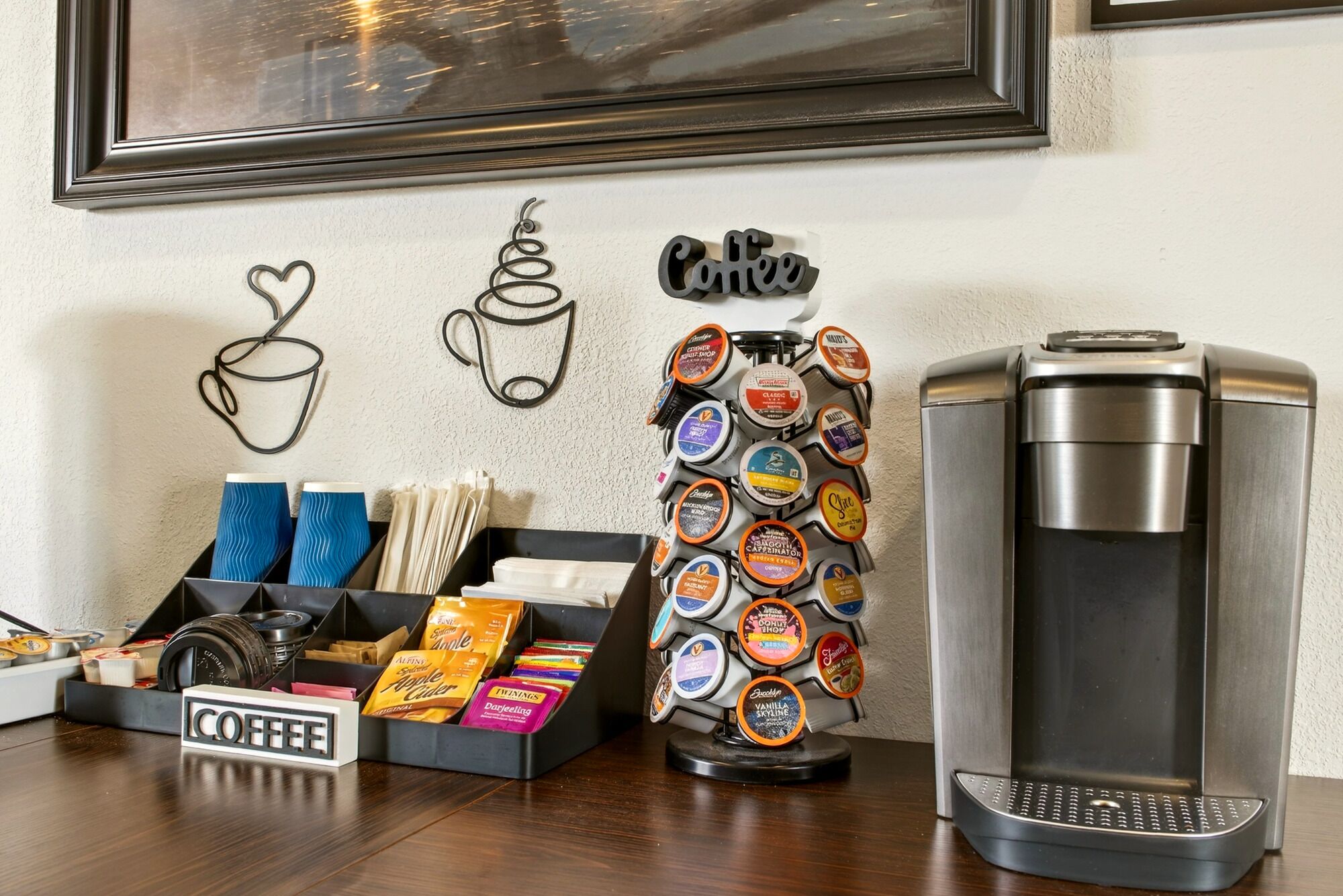 Una estación de café con una cafetera, un cuadro decorativo con motivos de café y un portalapilas para tazas de café.