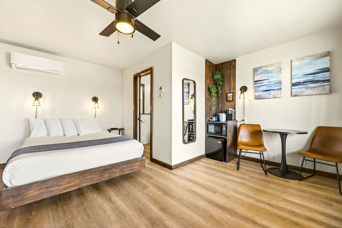 A room with a bed, a desk, two chairs, a ceiling fan, wall-mounted lights, a microwave, a coffee machine, and artwork.