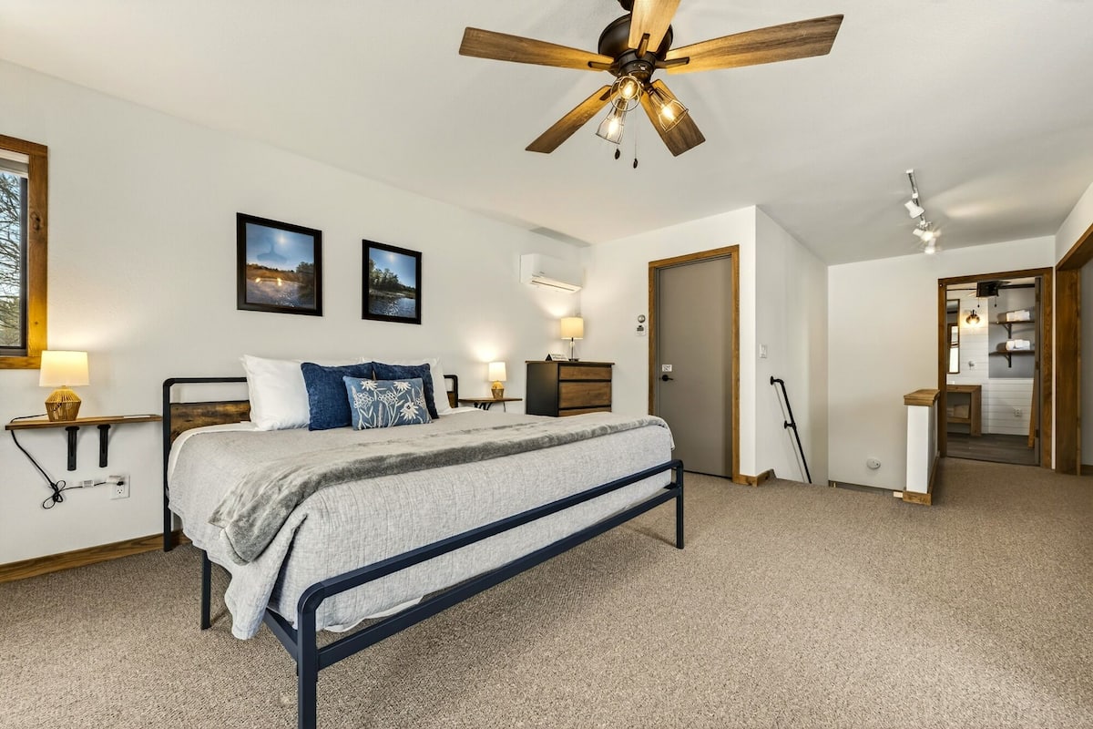 A bedroom with a bed, a ceiling fan, two wall-mounted picture frames, a bedside table with a lamp, and a door leading to another room.