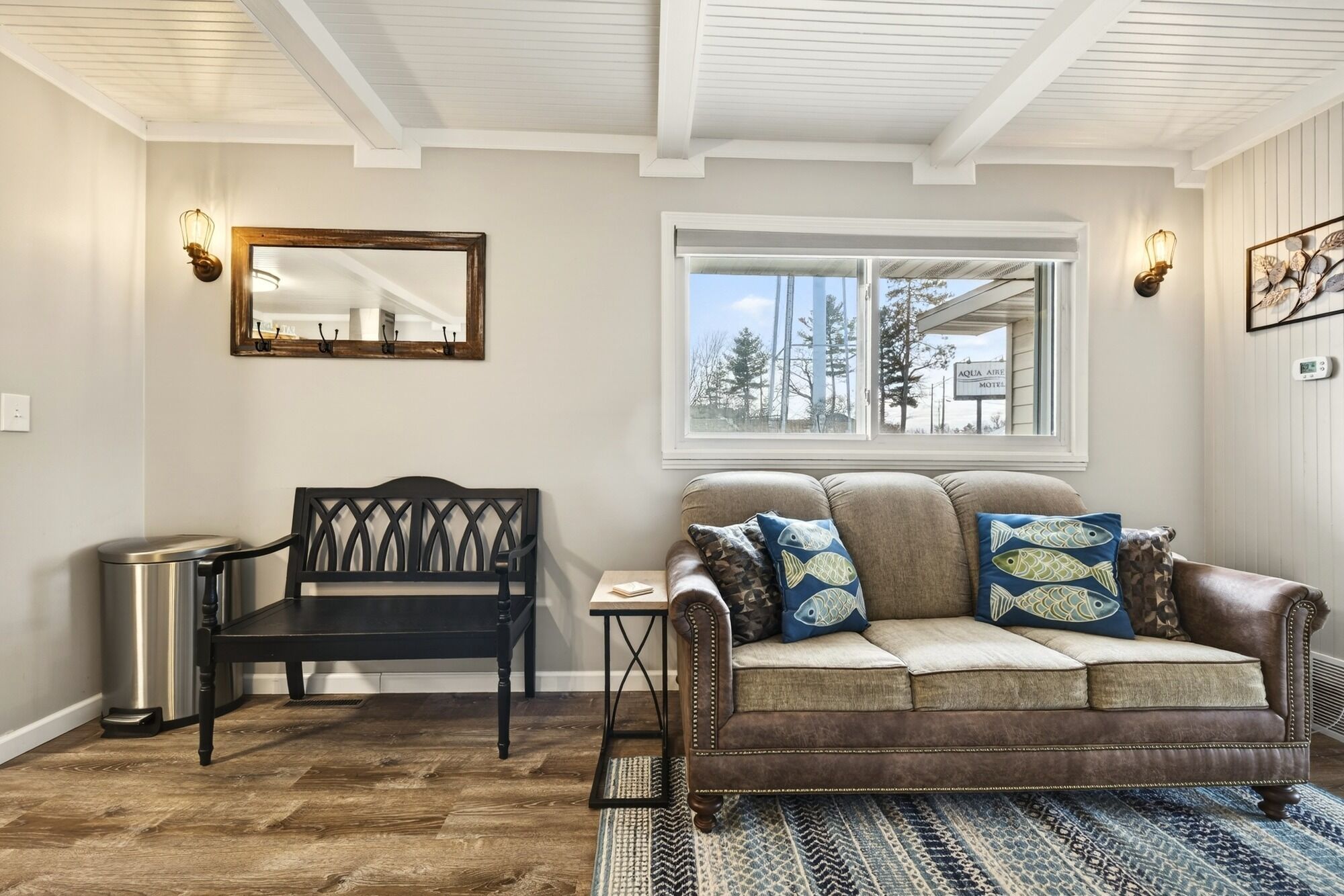 A living room with a brown leather sofa, a wooden bench, a mirror, and a window with a view of trees and a motel sign.