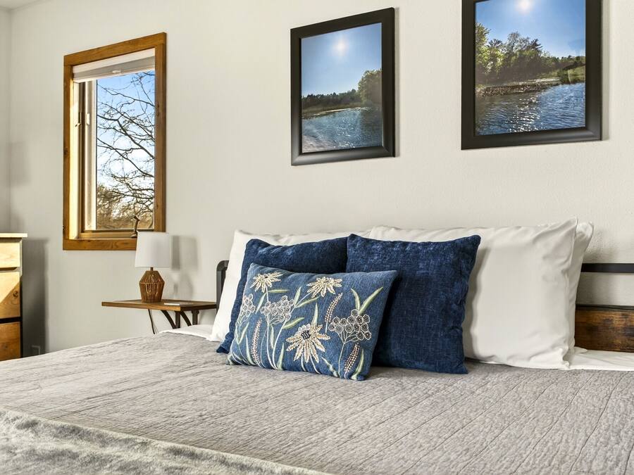 Un dormitorio con una cama, almohadas, una mesita de noche, una lámpara y cuadros en la pared.