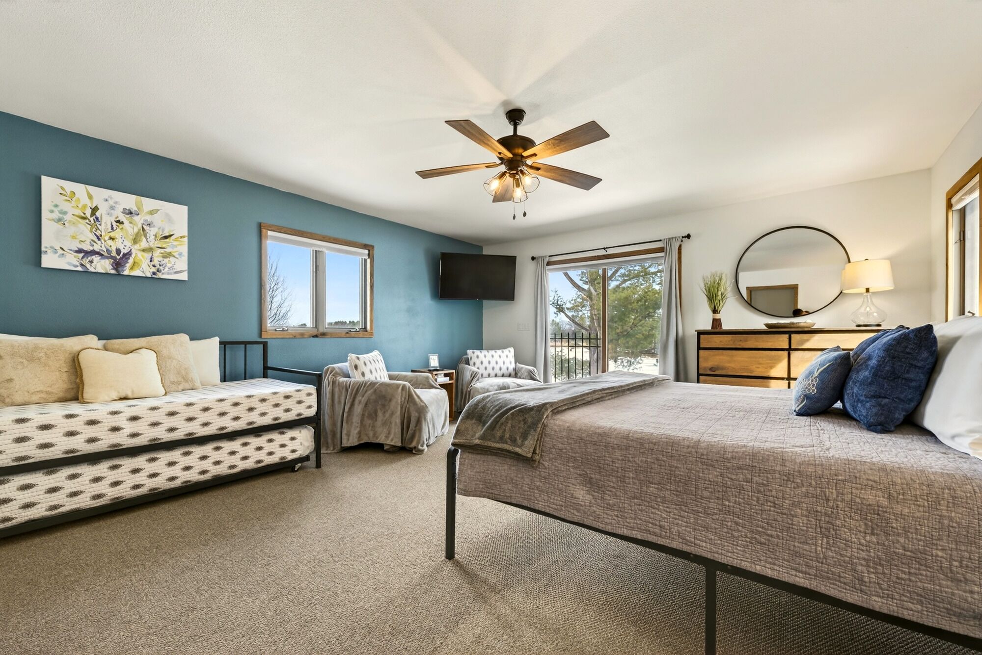 A bedroom with a bed, a sofa, a chair, a ceiling fan, a TV, a window, and a mirror.