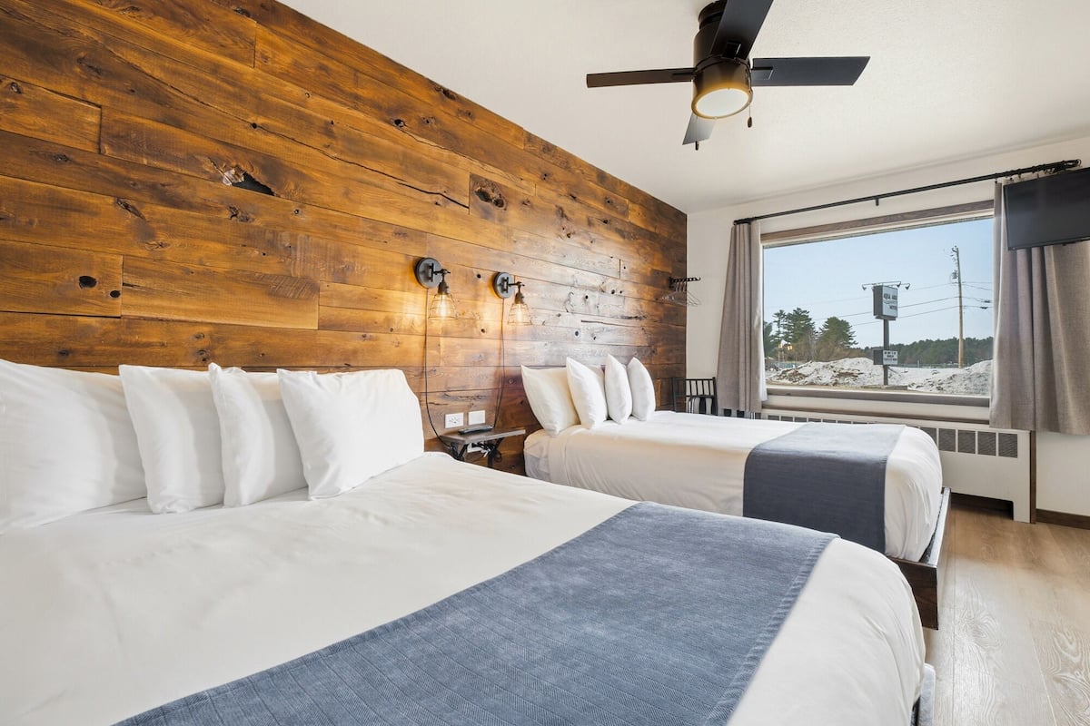 A hotel room with two beds, wooden headboard, ceiling fan, large window with view, and a nightstand with lamp.