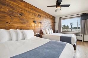 Quarto de hotel com duas camas, cabeceira de madeira, ventilador de teto, janela grande com vista e mesa de cabeceira com abajur.