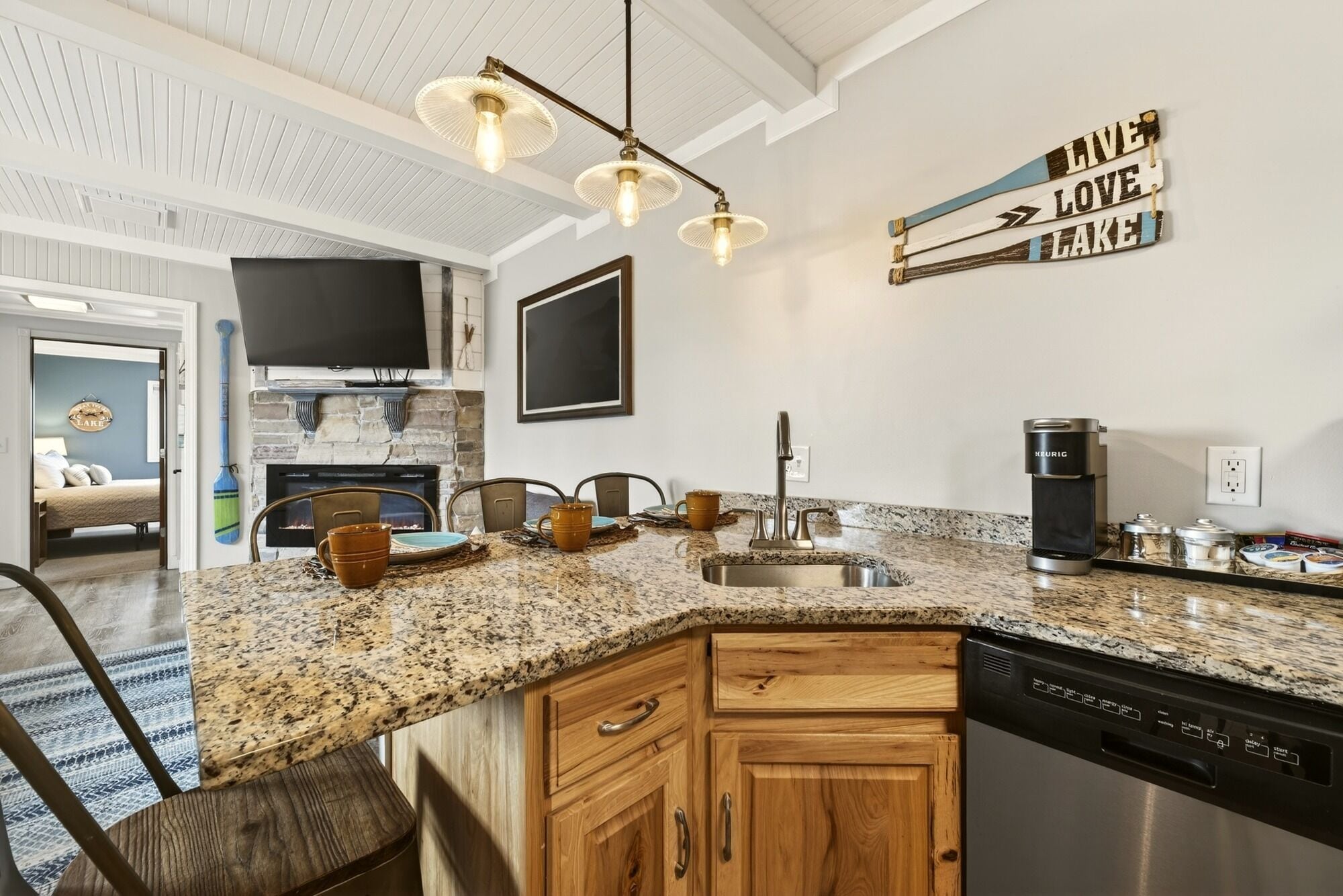 A kitchen with granite countertops, wooden cabinets, a dishwasher, a coffee maker, and a wall-mounted sign.