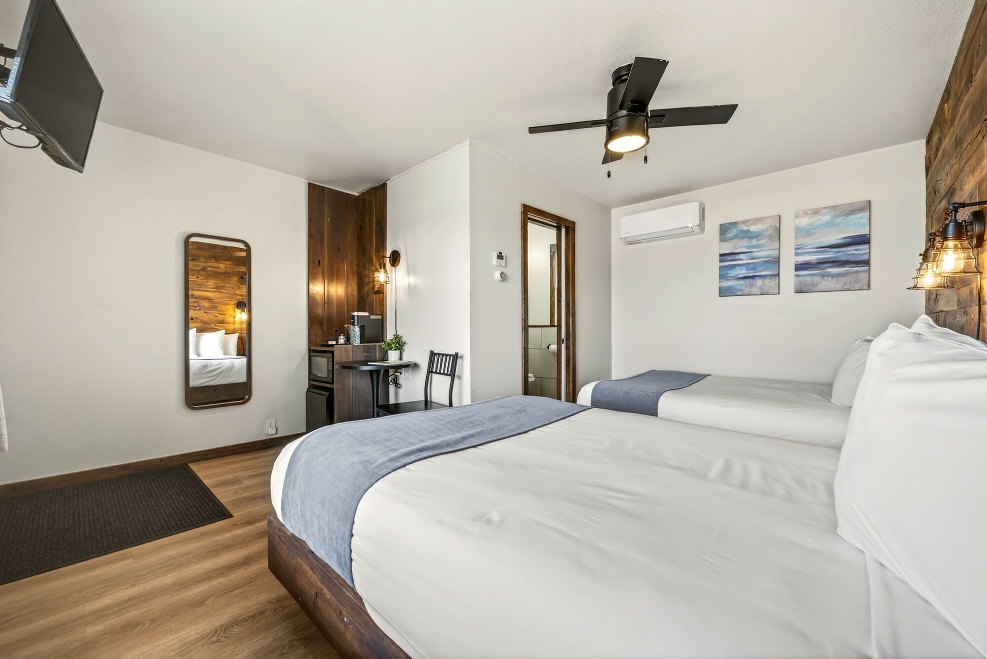 Quarto de hotel com cama, escrivaninha com cadeira, ventoinha no teto e obras de arte na parede.