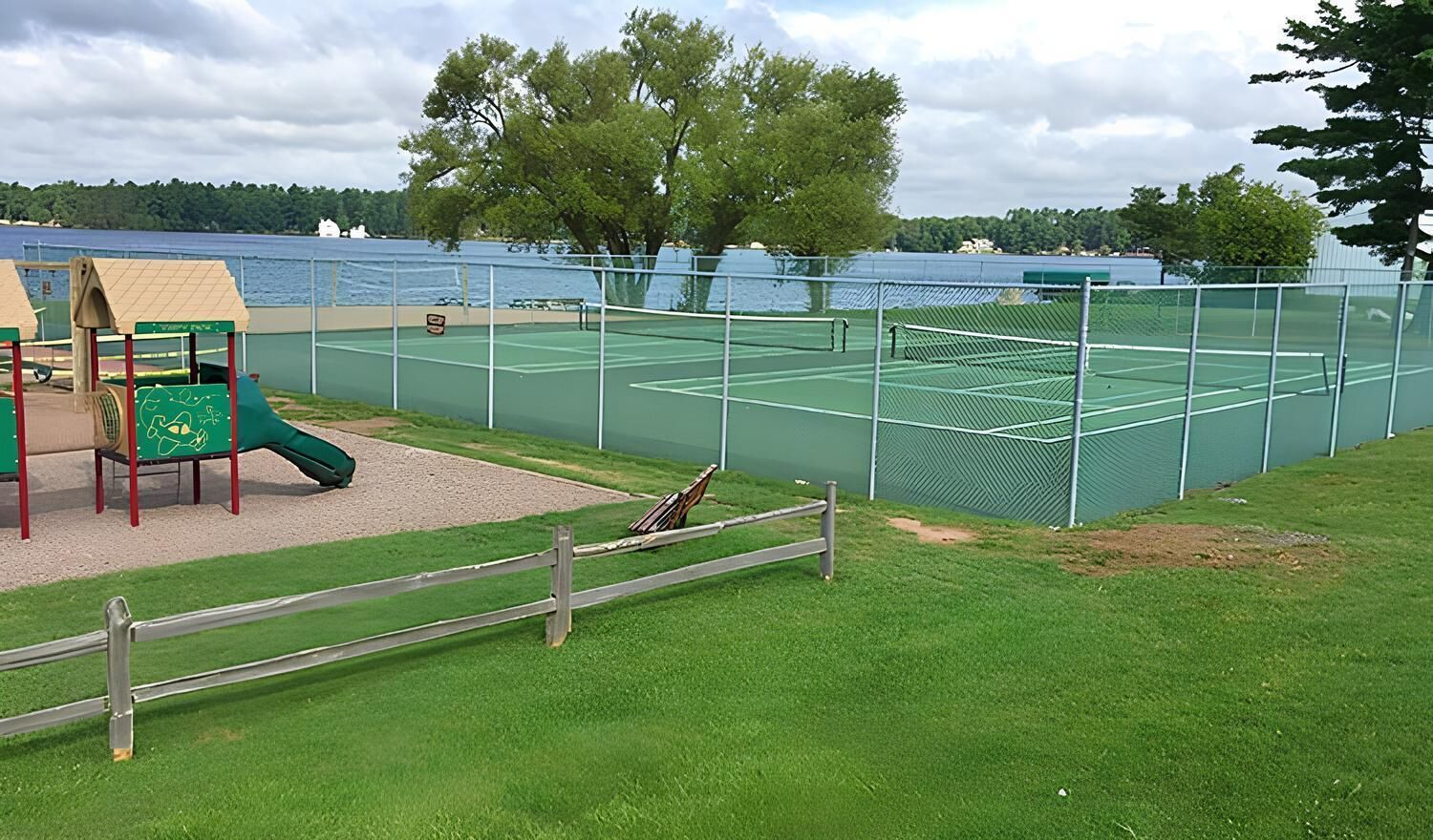 Una cancha de tenis con una red, rodeada por una valla, y un área de juegos con un tobogán y una estructura para escalar.