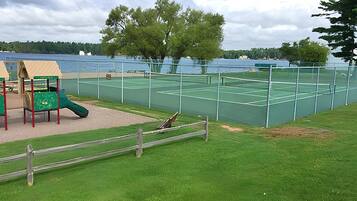 En tennisbane med net, omgivet af et hegn, samt en legeplads med rutschebane og klatrestruktur.