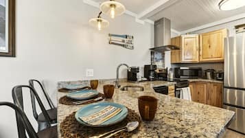 A kitchen with granite countertops, a stainless steel refrigerator, wooden cabinets, and a dining area with chairs and plates.