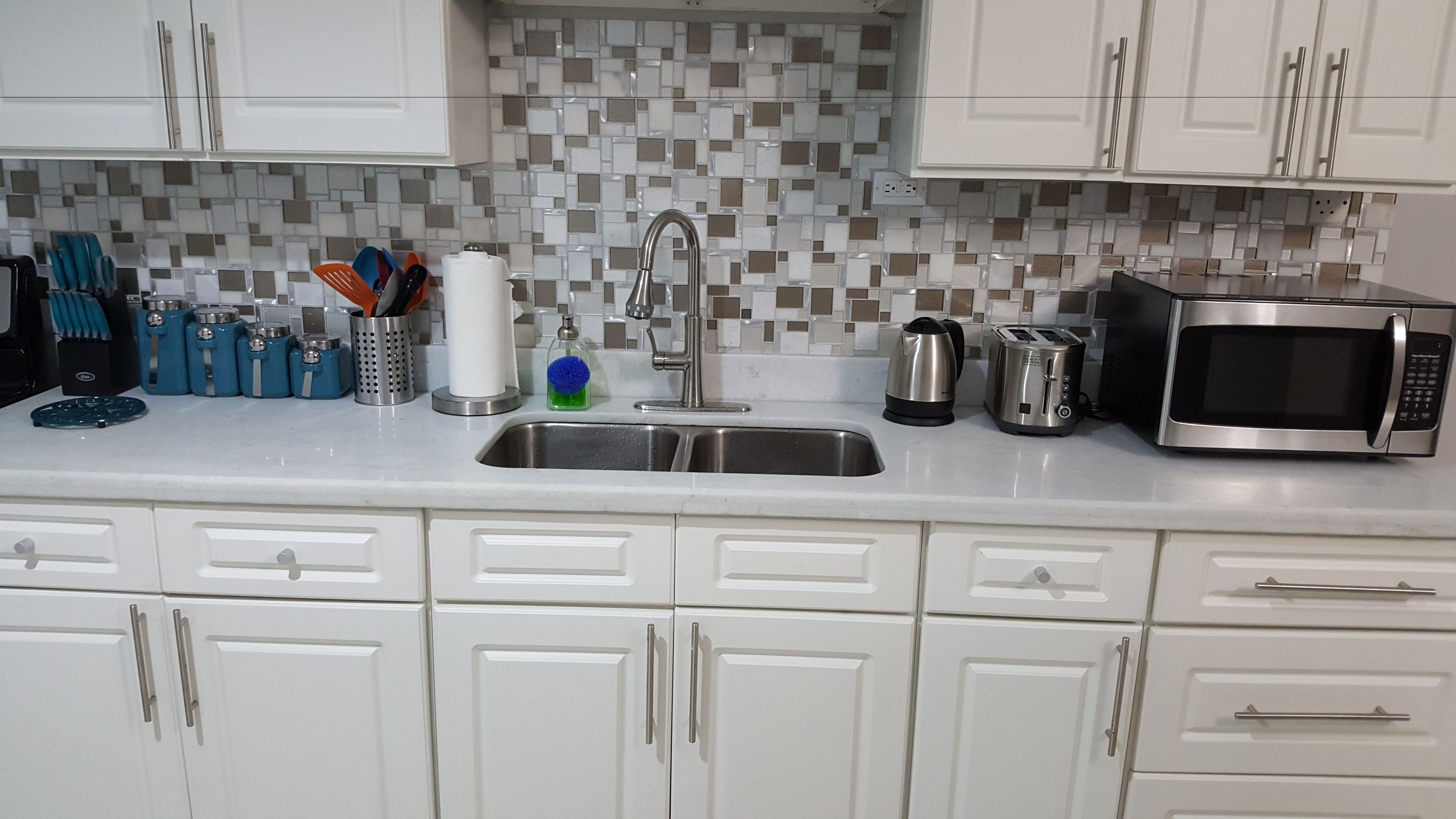 Cozinha com armários brancos, pia de aço inoxidável, chaleira, torradeira e micro-ondas.
