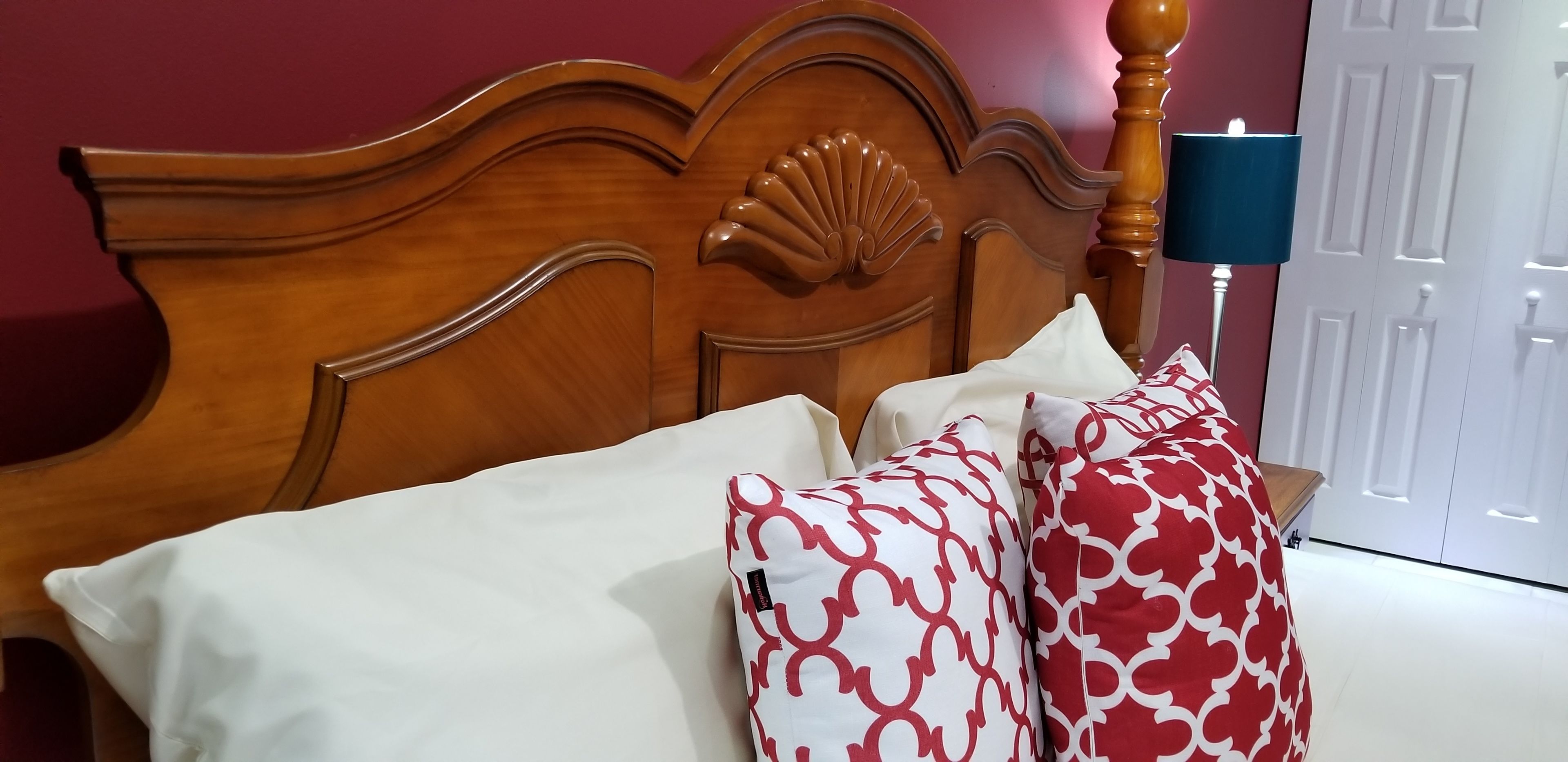 Un lit avec une tête de lit en bois, des oreillers blancs et rouges à motifs et une lampe de chevet bleue.