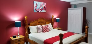 Un lit bien fait, recouvert d’une couverture rouge et blanche, deux lampes de chevet, un téléphone intelligent posé sur une table de nuit et un tableau accroché au mur.