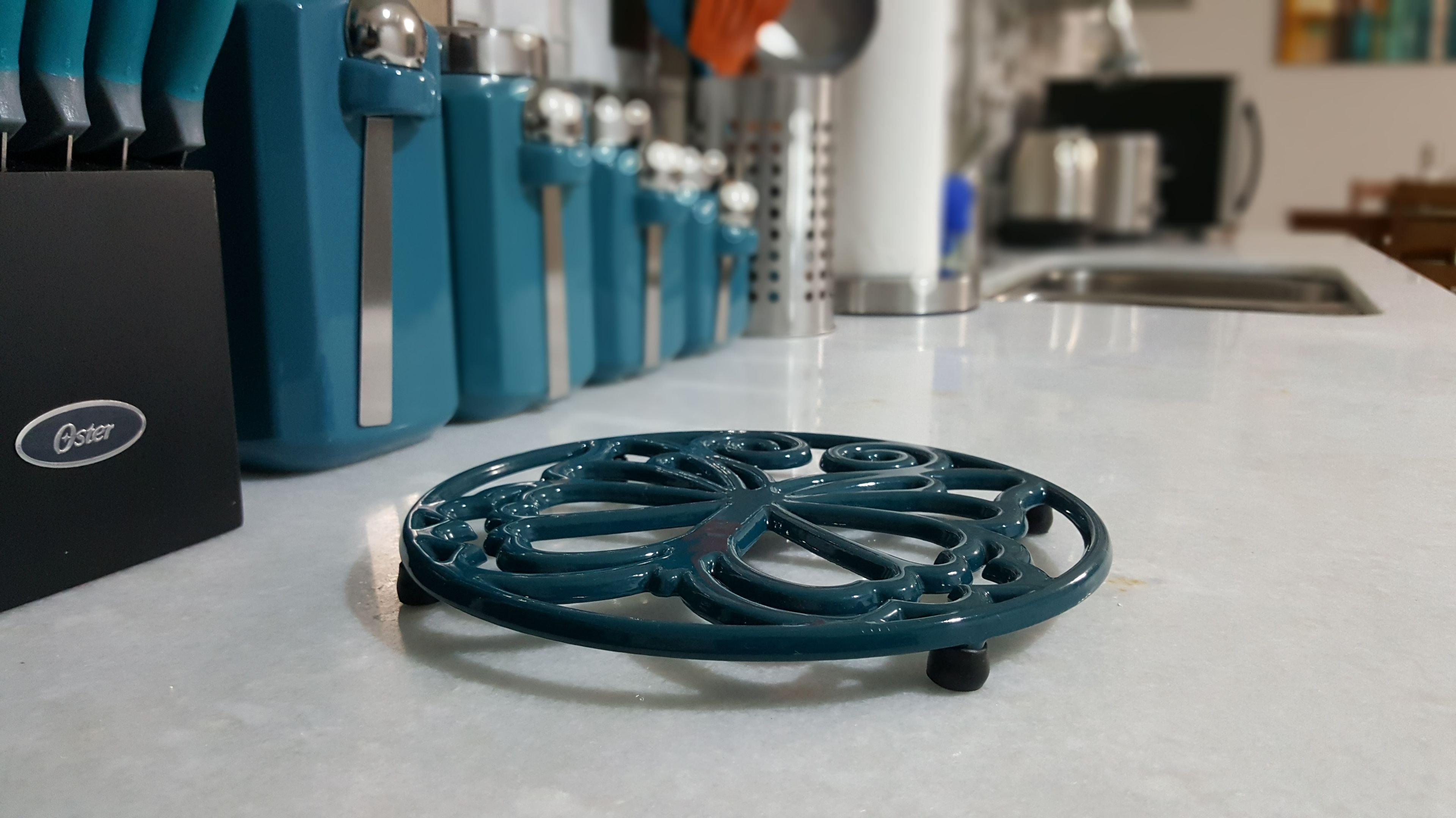 A kitchen counter with a blue appliance, a black box labeled "Oster," and a teal kitchen tool.
