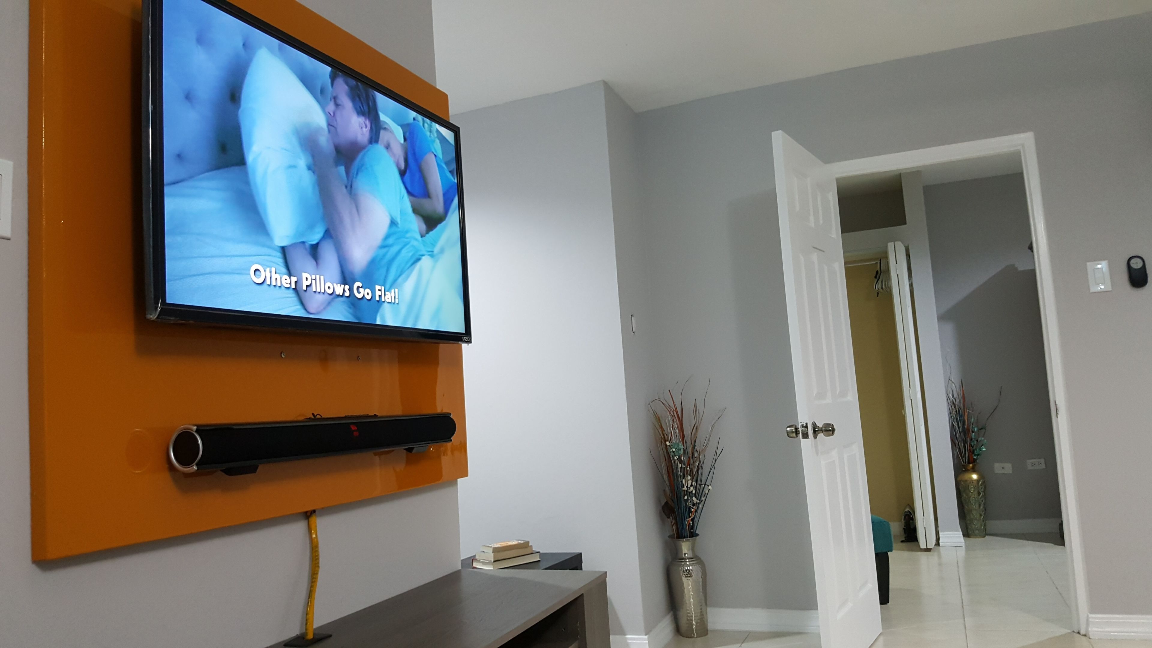 A flat-screen TV mounted on an orange wall, a white door leading to another room, and a vase with dried plants on the floor.