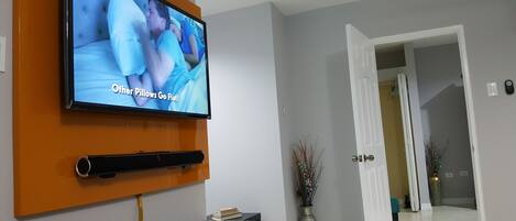 A flat-screen TV mounted on an orange wall, a white door leading to another room, and a vase with dried plants on the floor.
