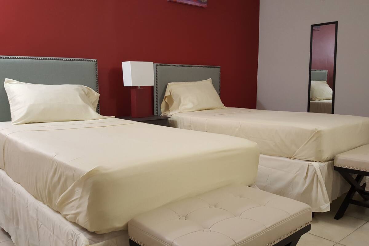 Une chambre d’hôtel avec deux lits, un banc, une table de chevet, une lampe et un miroir.