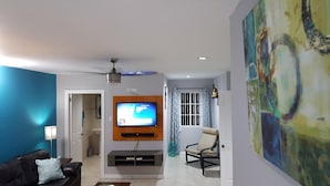 Una sala de estar con televisor de pantalla plana montado en la pared, ventilador de techo, sofá, mesa de centro y un cuadro en la pared.