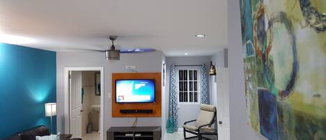 A living room with a flat-screen TV mounted on the wall, a ceiling fan, a sofa, a coffee table, and a painting on the wall.