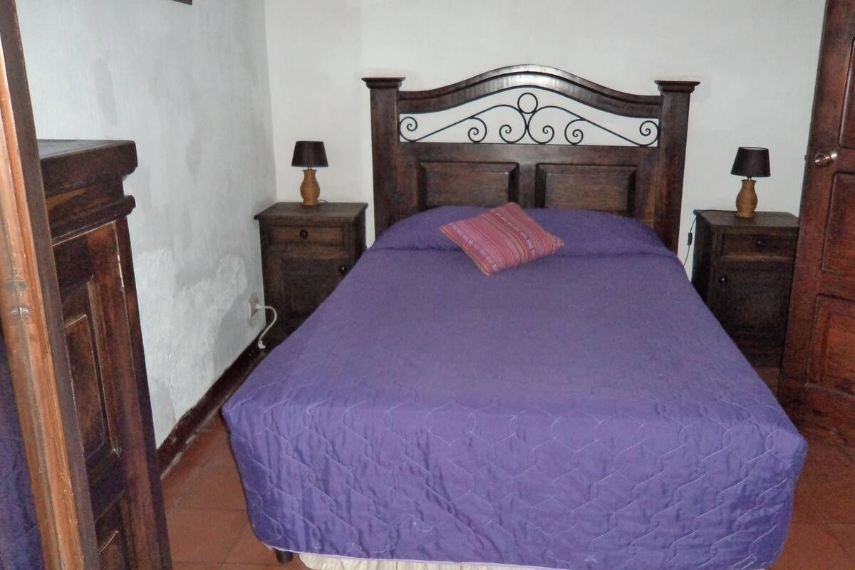 Un dormitorio con cabecera de madera, colcha morada y dos mesitas de noche con lámparas.