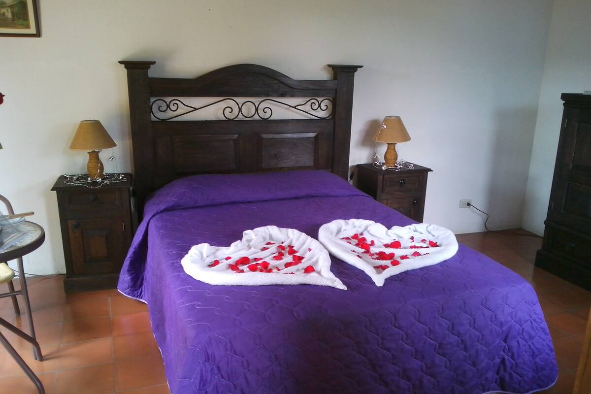 Una cama con un edredón morado y toallas con forma de corazón, adornada con pétalos de rosa.