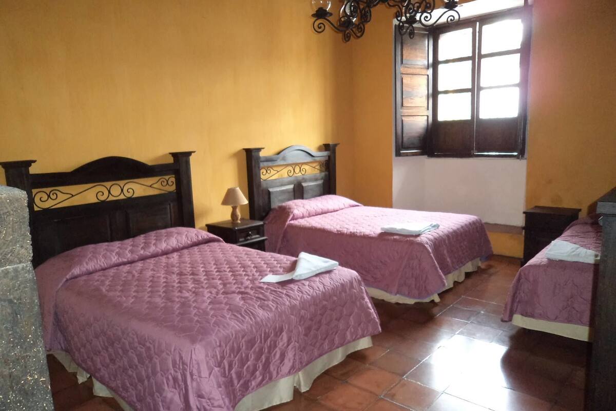 Una habitación con dos camas, cada una con cabecero de madera y colcha morada. Hay una ventana con persianas, una lámpara en una mesita de noche y un candelabro.