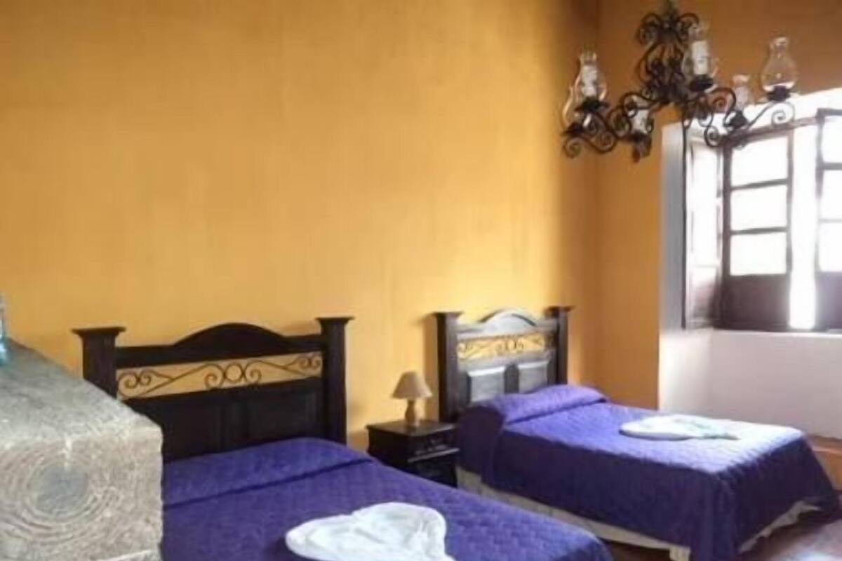 Una habitación con dos camas, cada una con una colcha morada y una toalla blanca sobre la cama. Entre las camas hay una mesita de noche con una lámpara. La pared es amarilla y hay una ventana con cortinas.
