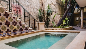 Uma piscina pequena com borda azulejada, uma escadaria com azulejos decorativos e uma parede de pedra ao fundo.