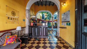 Um saguão de hotel com piso quadriculado, balcão de recepção, um banco com almofada colorida e uma placa para 'KAVIA HOTELS'.