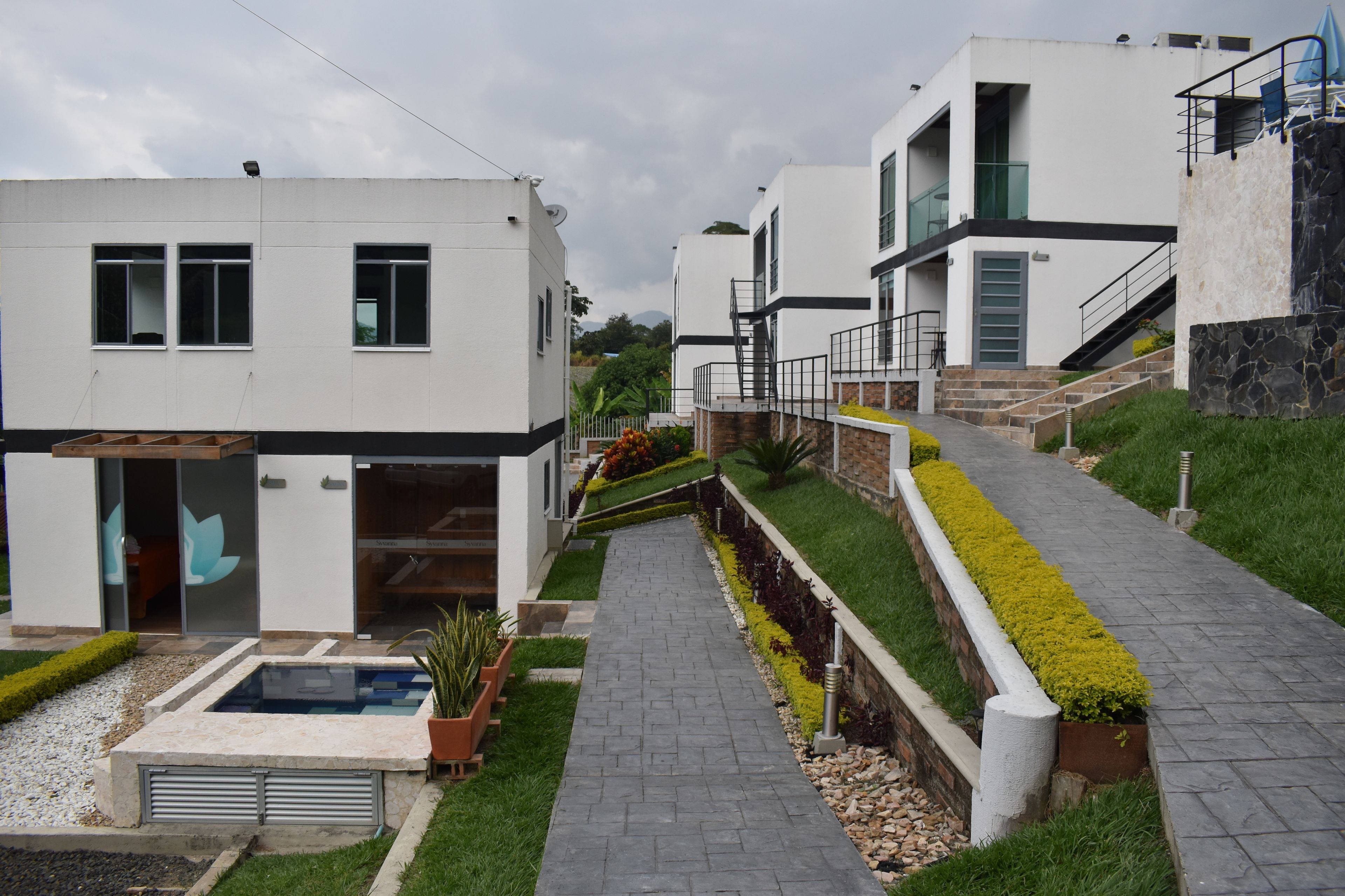 Un edificio residencial moderno con un sendero pavimentado, un pequeño jardín con fuente de agua y una escalera que conduce a un nivel superior.