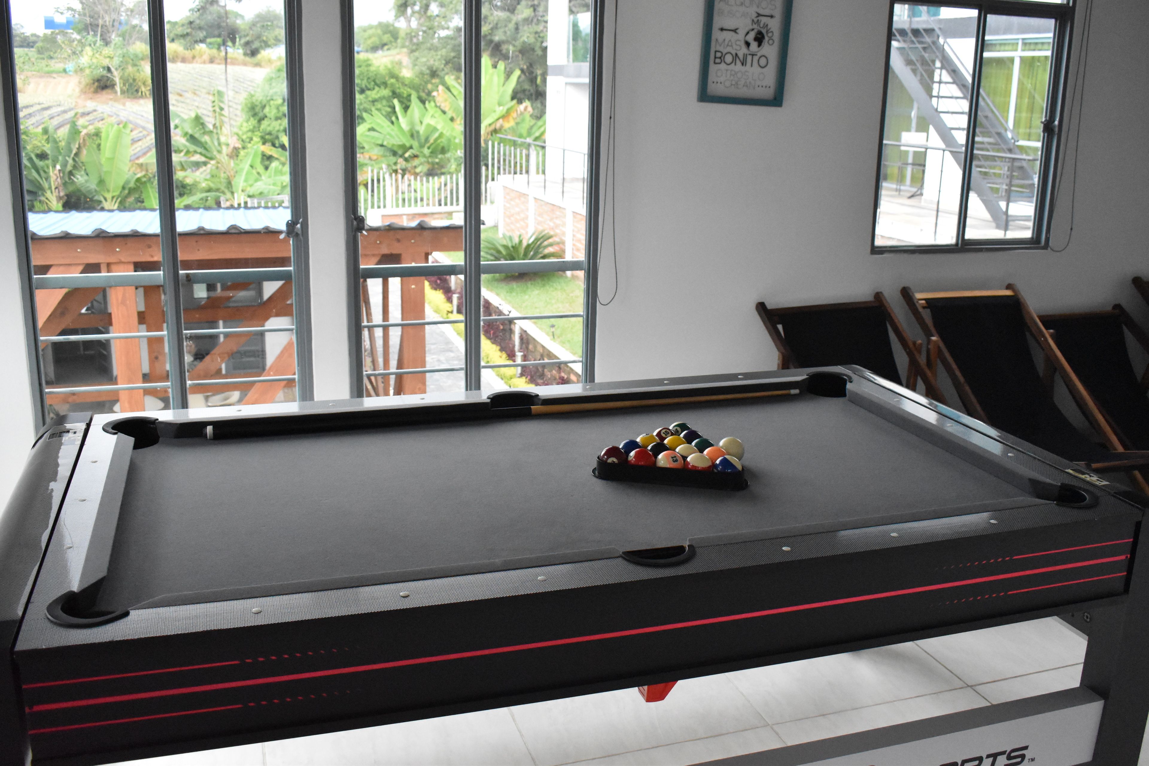 A pool table with balls, a window with a view of greenery, and a wall with a framed picture.