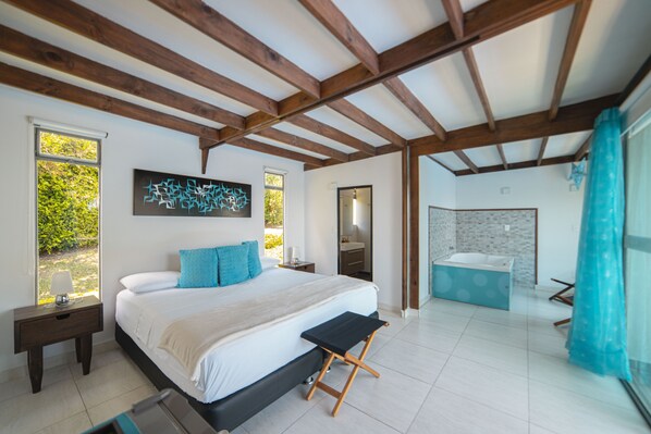 A bedroom with a large bed, a nightstand, a window with a view, a bathroom with a bathtub, and wooden beams on the ceiling. - Syvanna Hotel Wellness & SPA (Pereira)