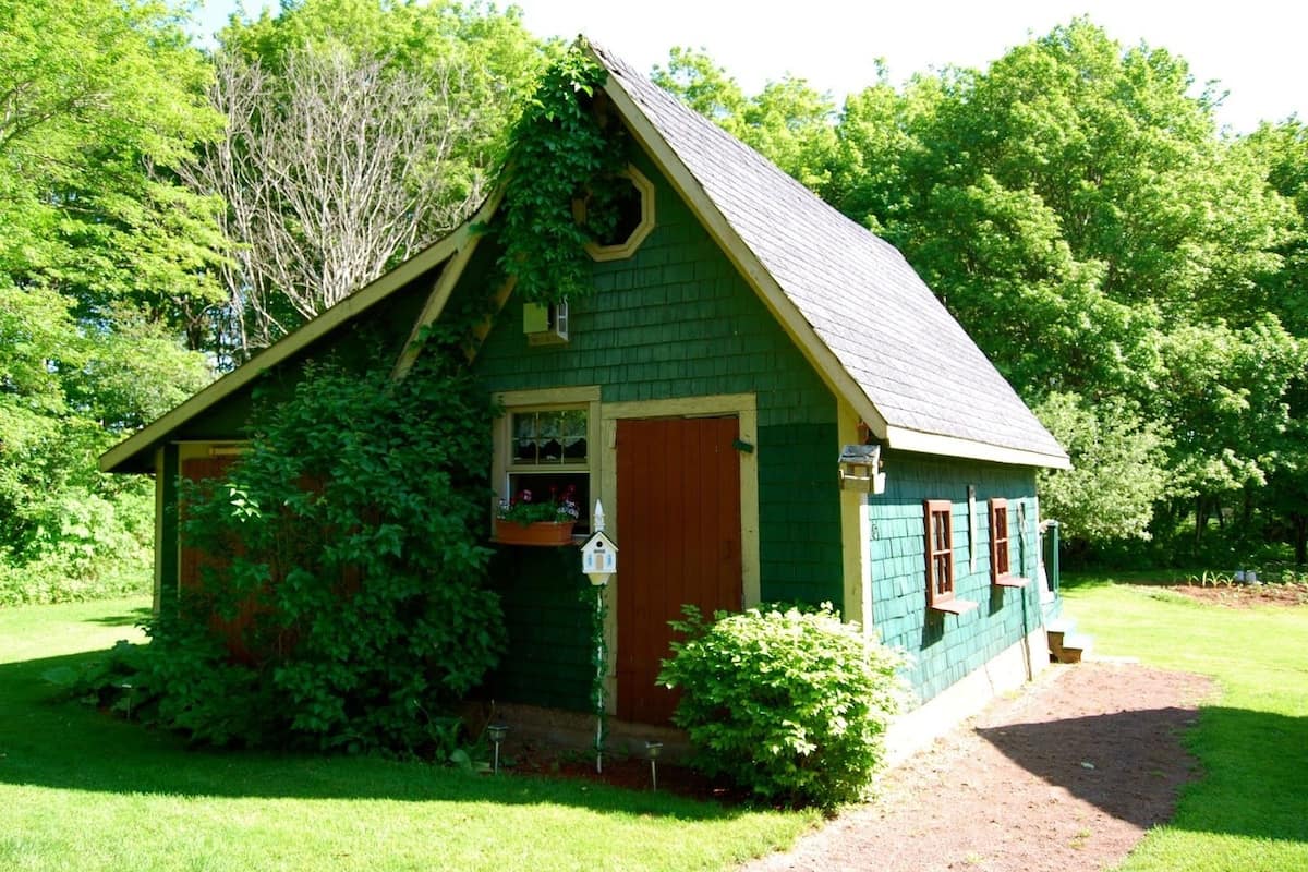 A small, green wooden house with a sloped roof, a front door, and a window with a hanging planter.