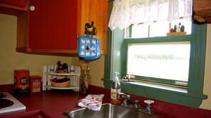 A kitchen with a stainless steel sink, a window with lace curtains, and a view of houses outside. - Bugaboo Cottages (Central Bedeque)